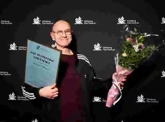 Regissären Mårten Nilsson poserar leende med ett diplom i ena handen och en bukett blommor i den andra handen