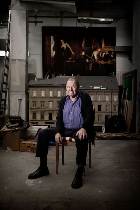 Roy Andersson sitter på en stol omgiven av scenografin från en av hans filmer