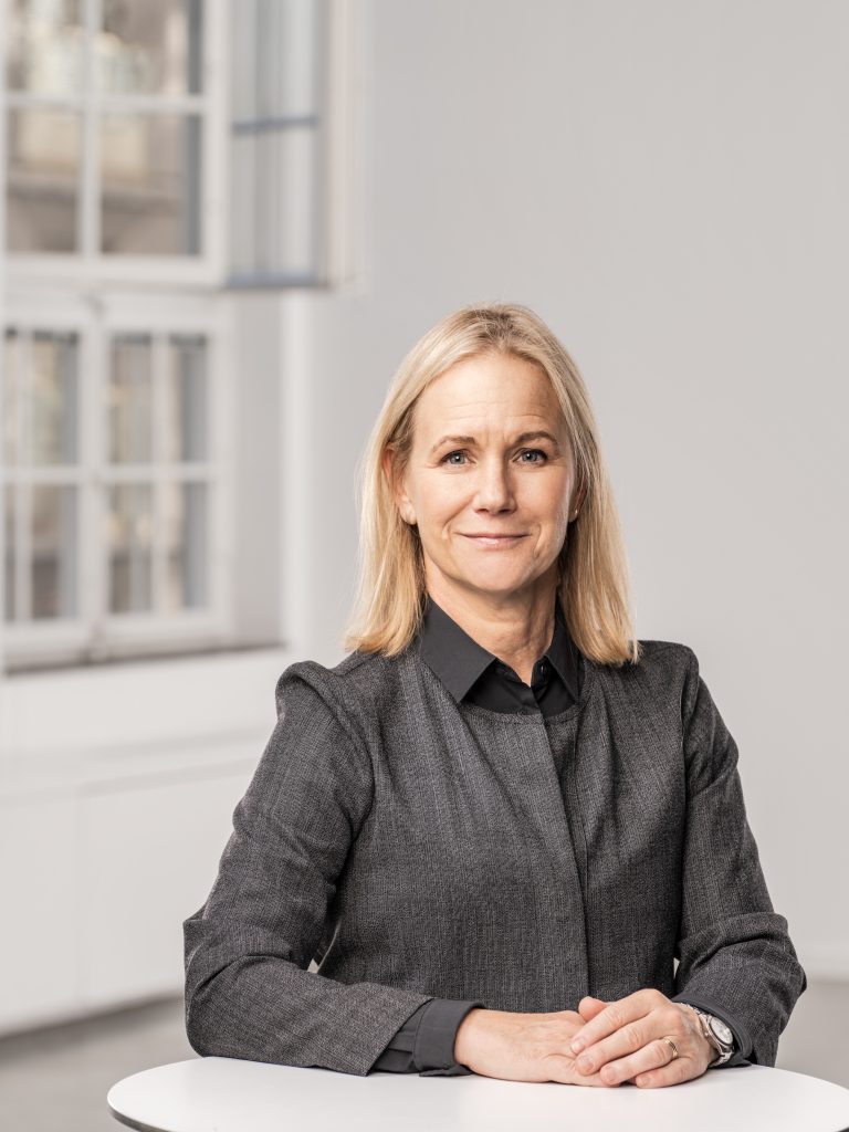 Mika står lätt vriden och tittar leende in i kameran. Man ser överkroppen och ett fönster i bakgrunden. Fotografiet är i färg.