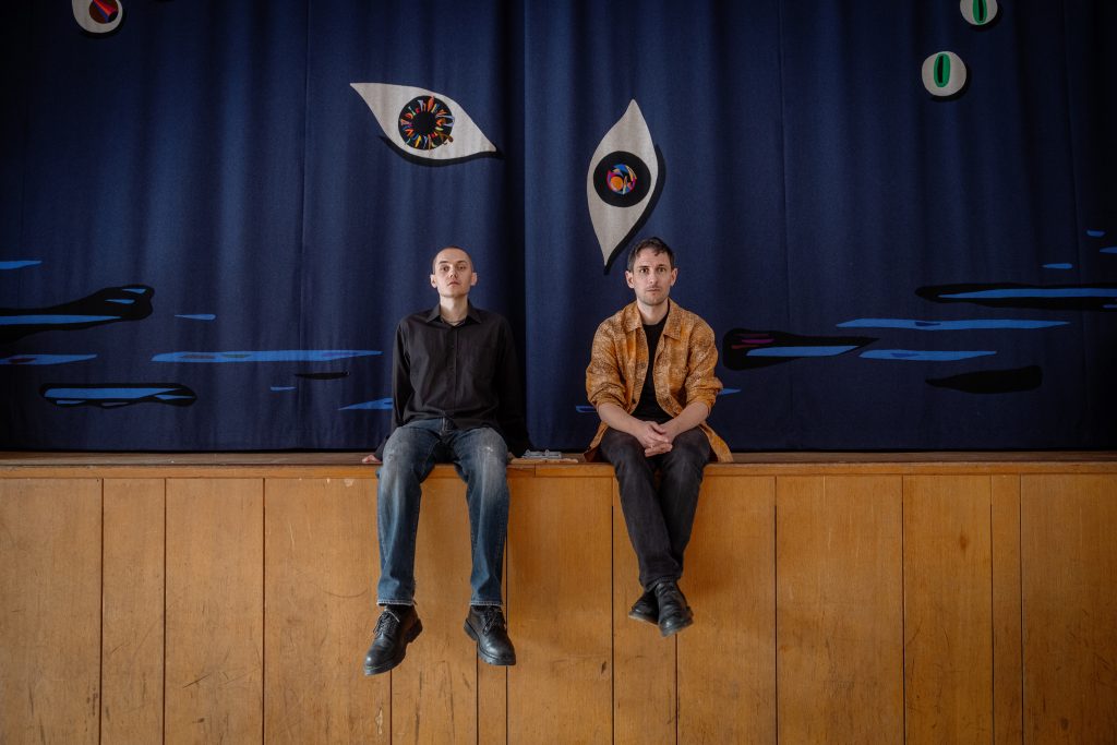 Sebastian Dahlqvist och Elof Hellström porträtterade framför ridån i Hägerstensåsens medborgarhus som är gjord av konstnären Laleh Kazemi Veisari 