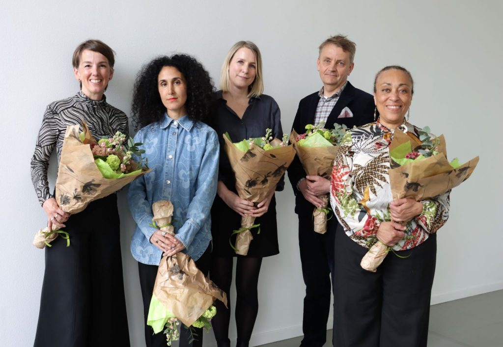 Gruppbild i färg på fem leende personer som poserar framför en ljus bakgrund och de håller i stora blombuketter.