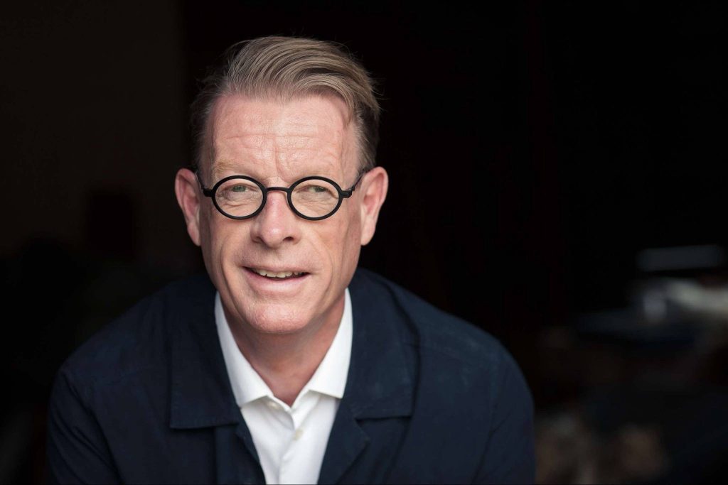 Närbild på en man - Benny Marcel som är Konstnärsnämndens nya ordförande, med glasögon, iklädd vit skjorta och mörk kavaj framför en mörk bakgrund.