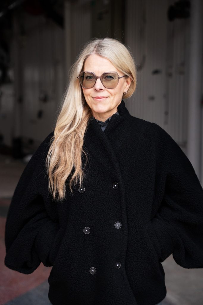 Porträttbild av Gunilla Klingberg, kvinna i långt blond hår, svart jacka och solglasögon.