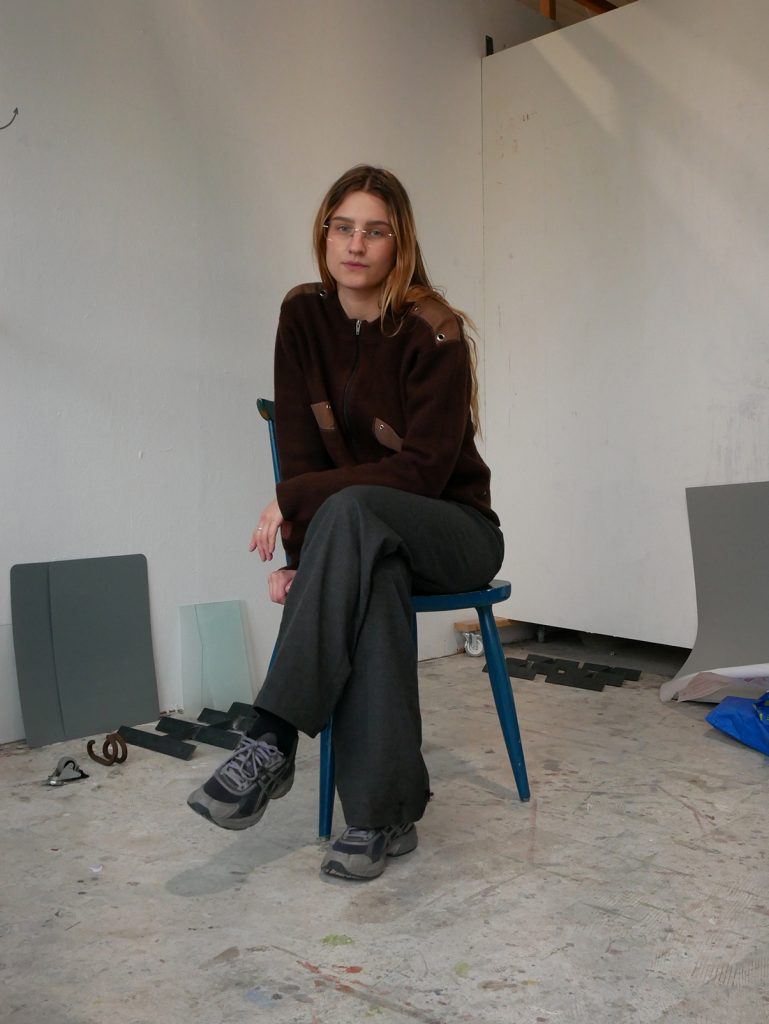 Konstnär Sara Andreasson sitter med benen i kors på en stol i en ateljé, hon har långt mellanbrunt hår och 