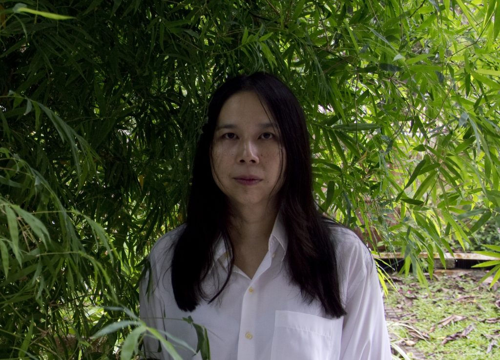 Portrait of the artist. Greeneries in the background. Kah Bee wears a white shirt and have long dark hair.