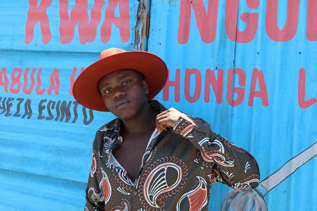 Tusse i mönstrad skjorta och färgglad hatt framför en blå vägg.